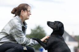 weibliche Person schaut grinsend ihren Hund an, der vor ihr sitzt und eine Pfote auf ihrem Arm hat, während sie seine Brust krault, fotografiert von LiraAmarokPictures