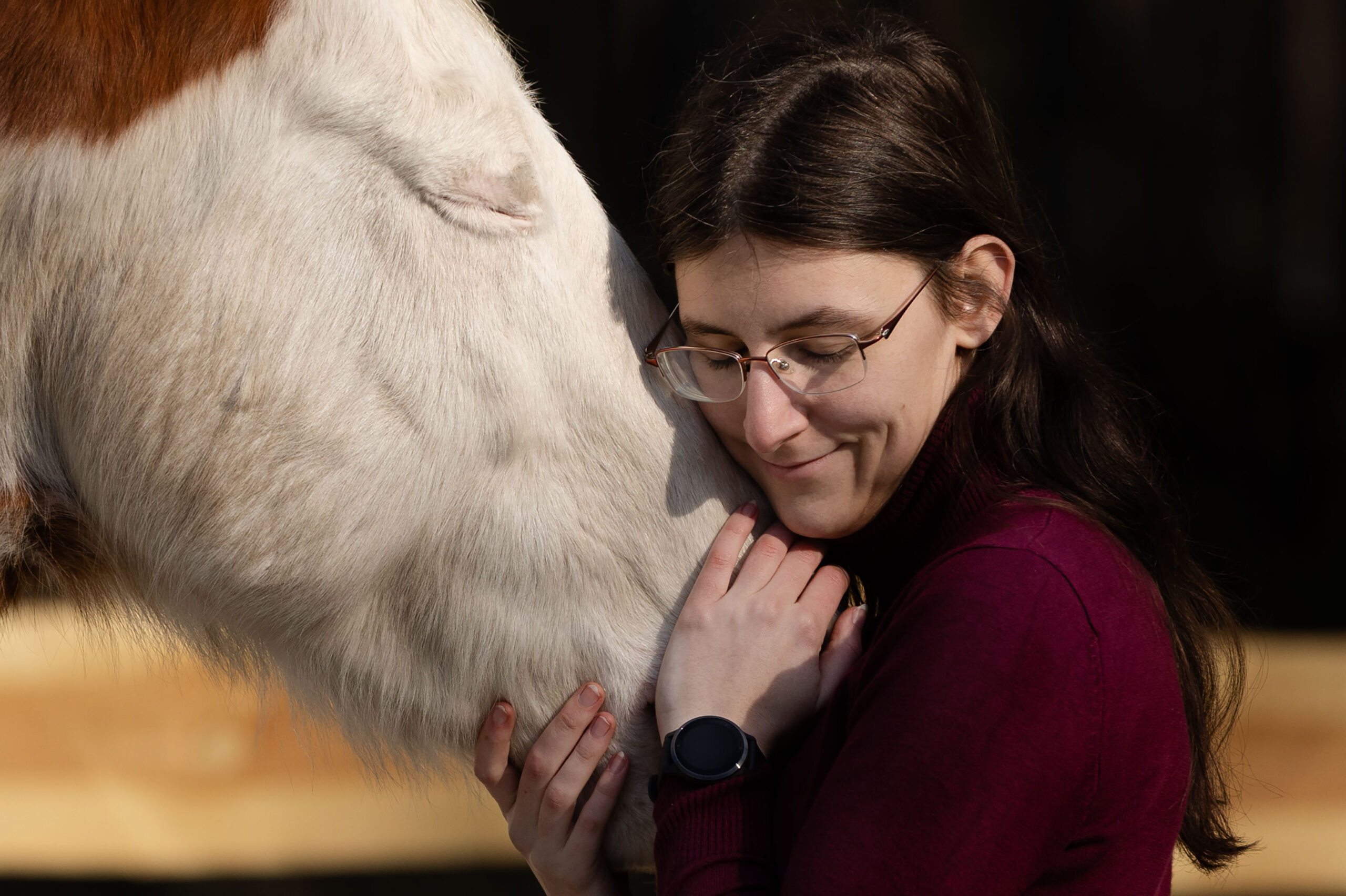 Frau mit braunen Haaren und Brille kuschelt sich an die Nüstern eines weiß-braun-gescheckten Pferdes. Beide haben die Augen zu