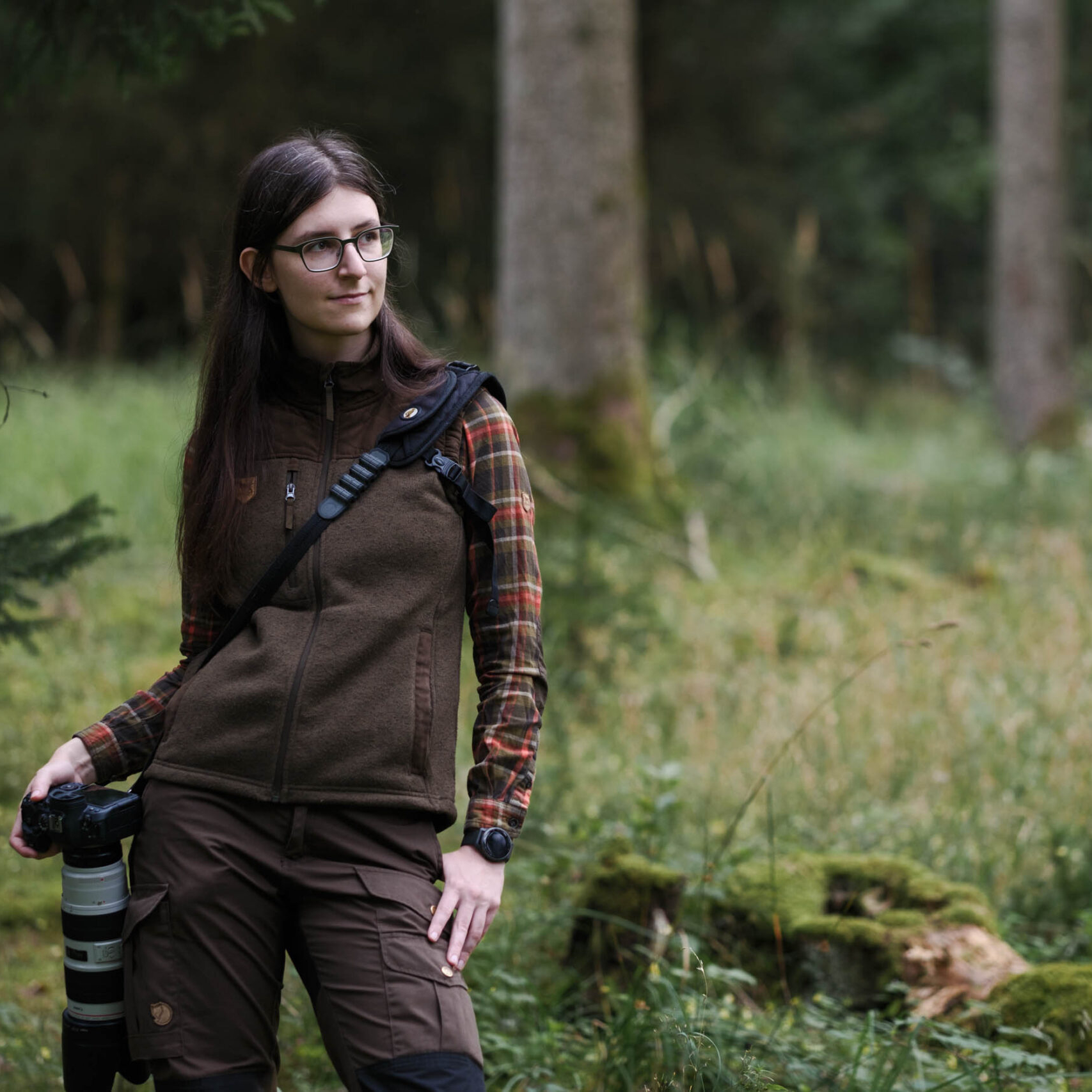 Bild von einer Frau (Lira) im Wald stehend. Sie trägt Jagdkleidung mit einer Weste und hat die Kamera an der Seite hängend. Sie blickt zur Seite.