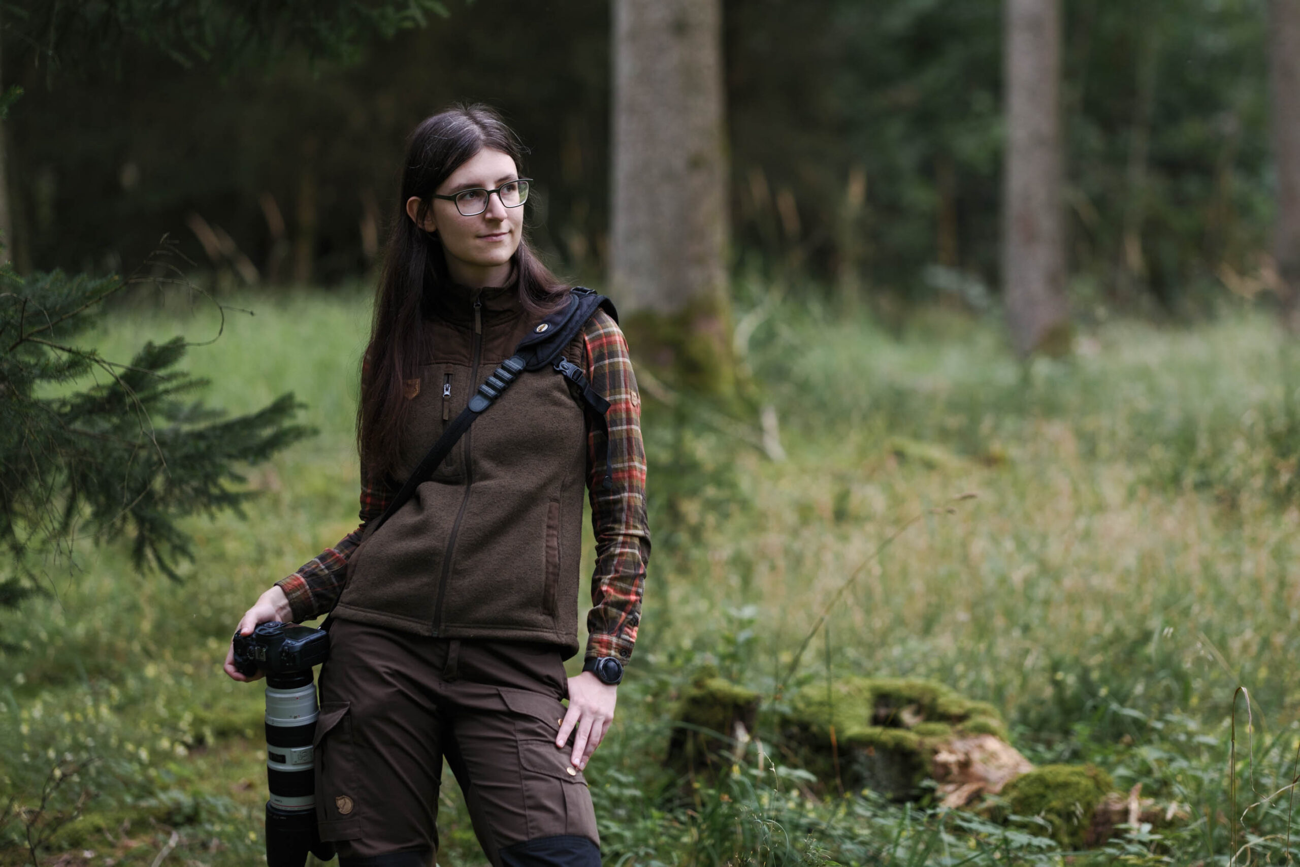 Bild von einer Frau (Lira) im Wald stehend. Sie trägt Jagdkleidung mit einer Weste und hat die Kamera an der Seite hängend. Sie blickt zur Seite.