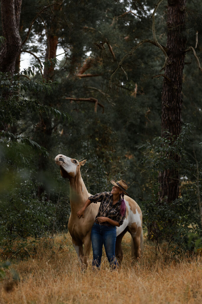 geschecktes Pferd steht mit einer Frau im Wald und hat den Kopf lang gestreckt, weil es an der Brust gekrault wird.