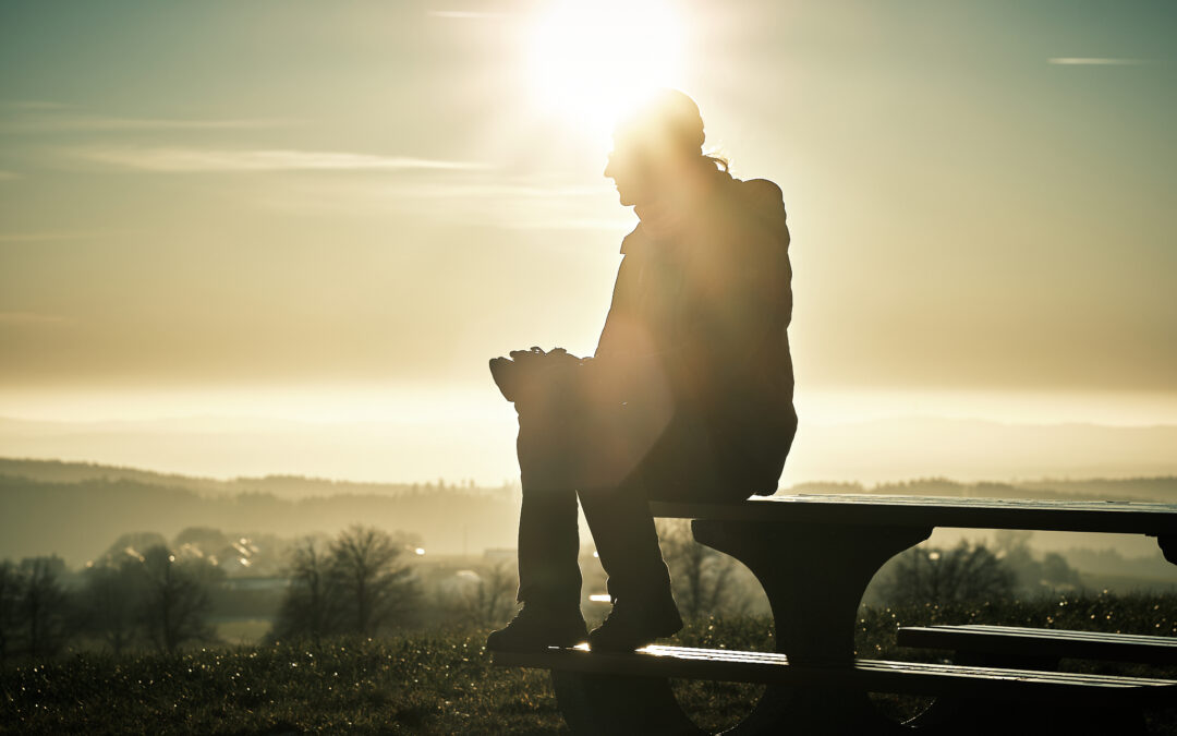 Silouette einer Frau auf einer Bank sitzend im Winter, hinter ihr steht die Sonne am Himmel