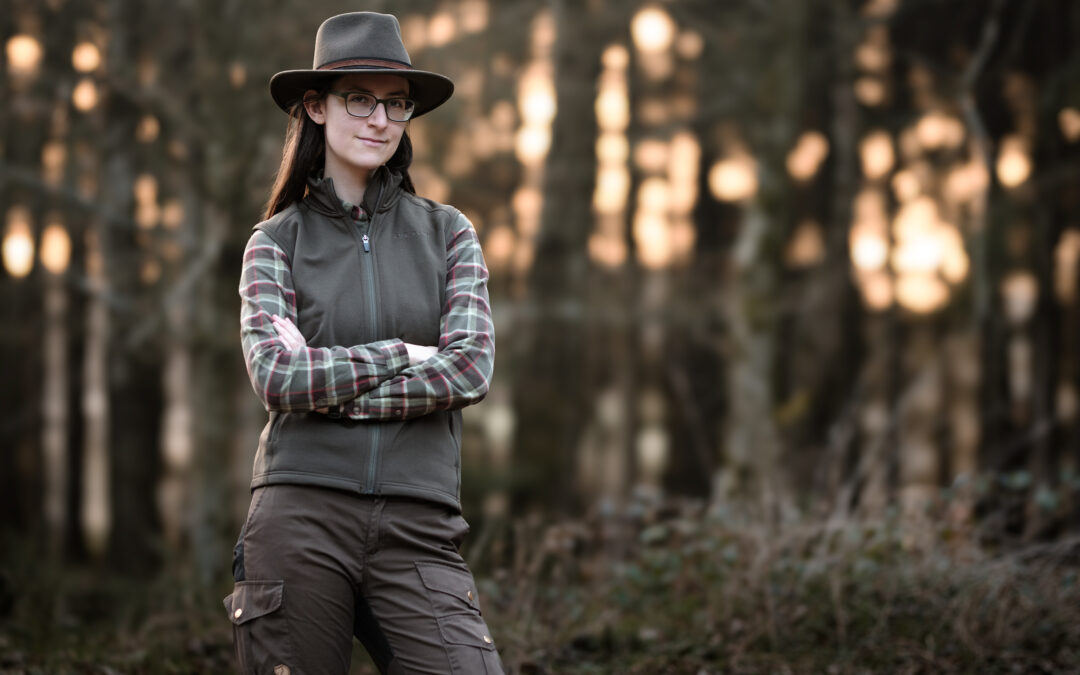 Eine Frau in Jagdkleidung mit Jagdhut steht mit verschränkten Armen im Wald