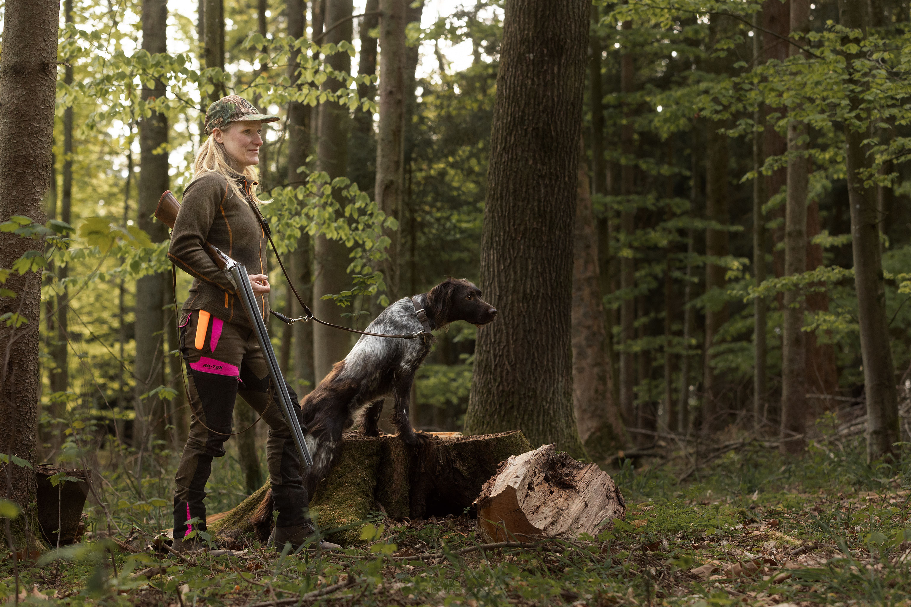 Jägerin mit gebrochener Flinte überm Arm steht mit ihrem Hund zusammen im Wald