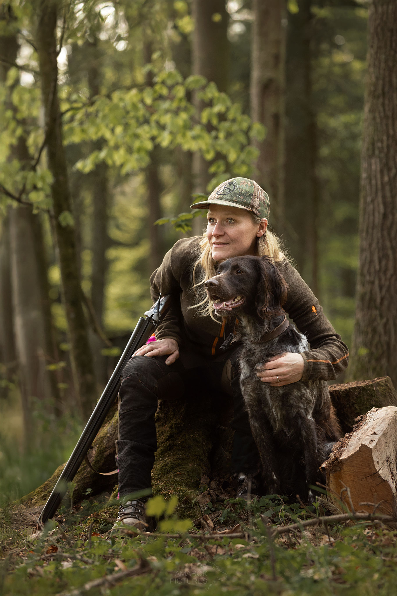 Jägerin mit Flinte überm Arm gebrochen sitzt auf einem Baumstumpf und blickt mit ihrem kleinen Münsterländer in die Ferne