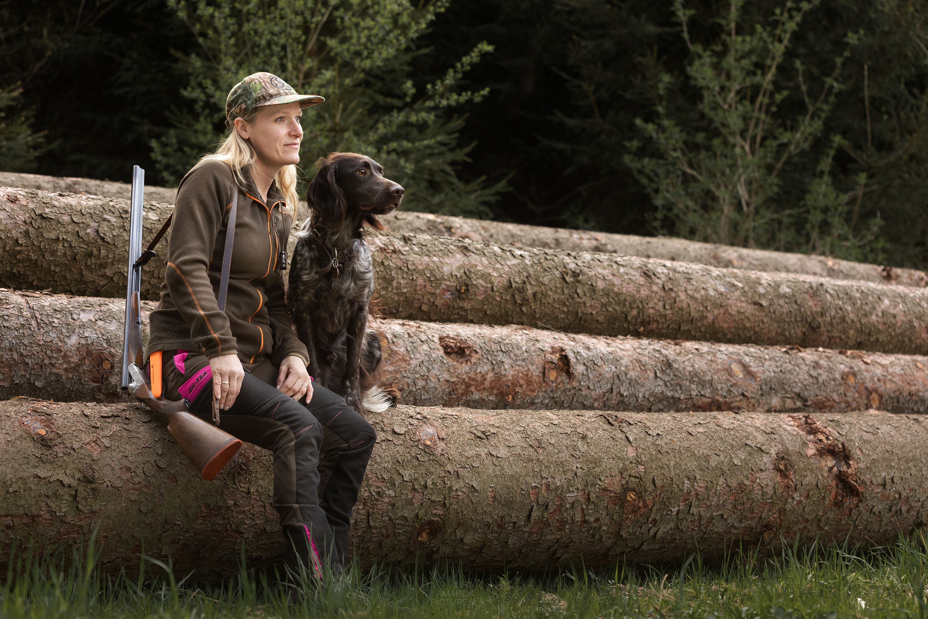 Eine Jägerin mit Flinte und ihrem kleinen Münsterländer sitzen auf einem Holzpolter und blicken in die Ferne