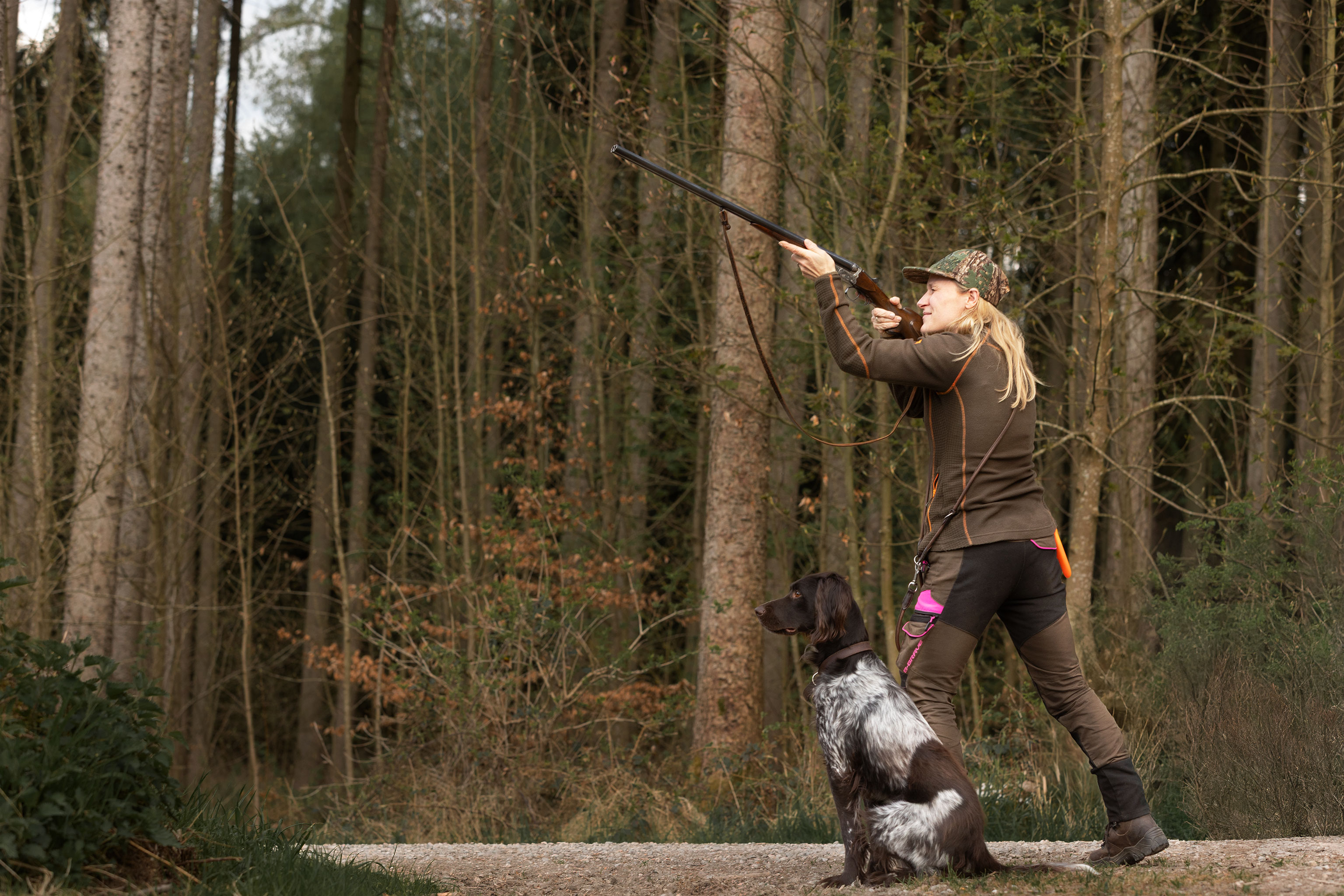 Eine Jägerin mit Flinte im Anschlag, ihr Hund sitzt neben ihr