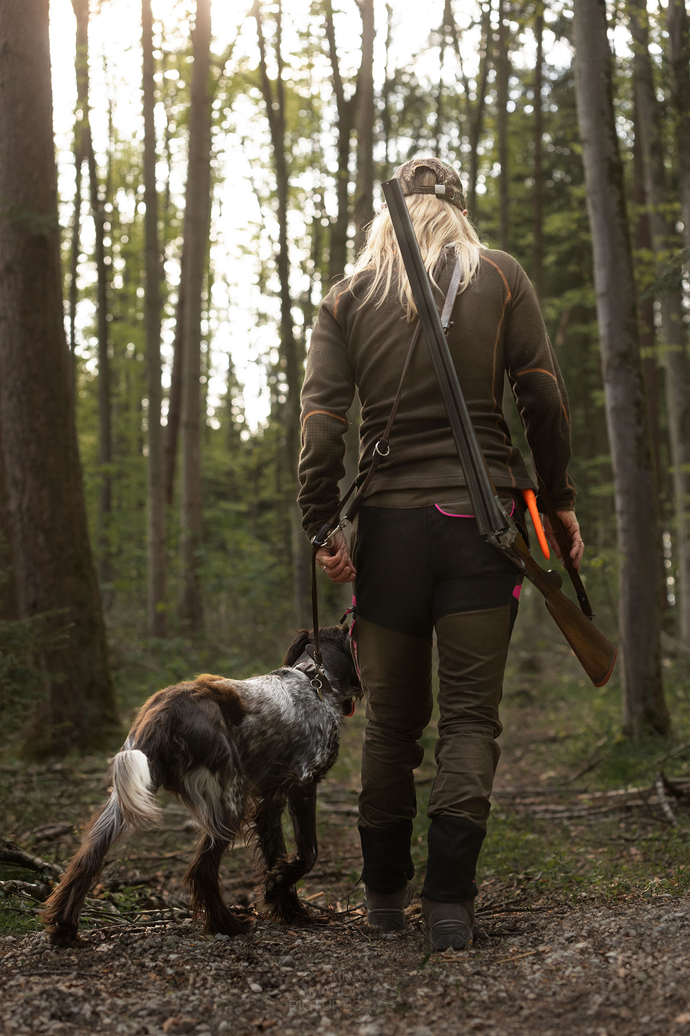 Eine Jägerin mit gebrochener Flinte über der Schulter geht mit ihrem Hund durch den Wald