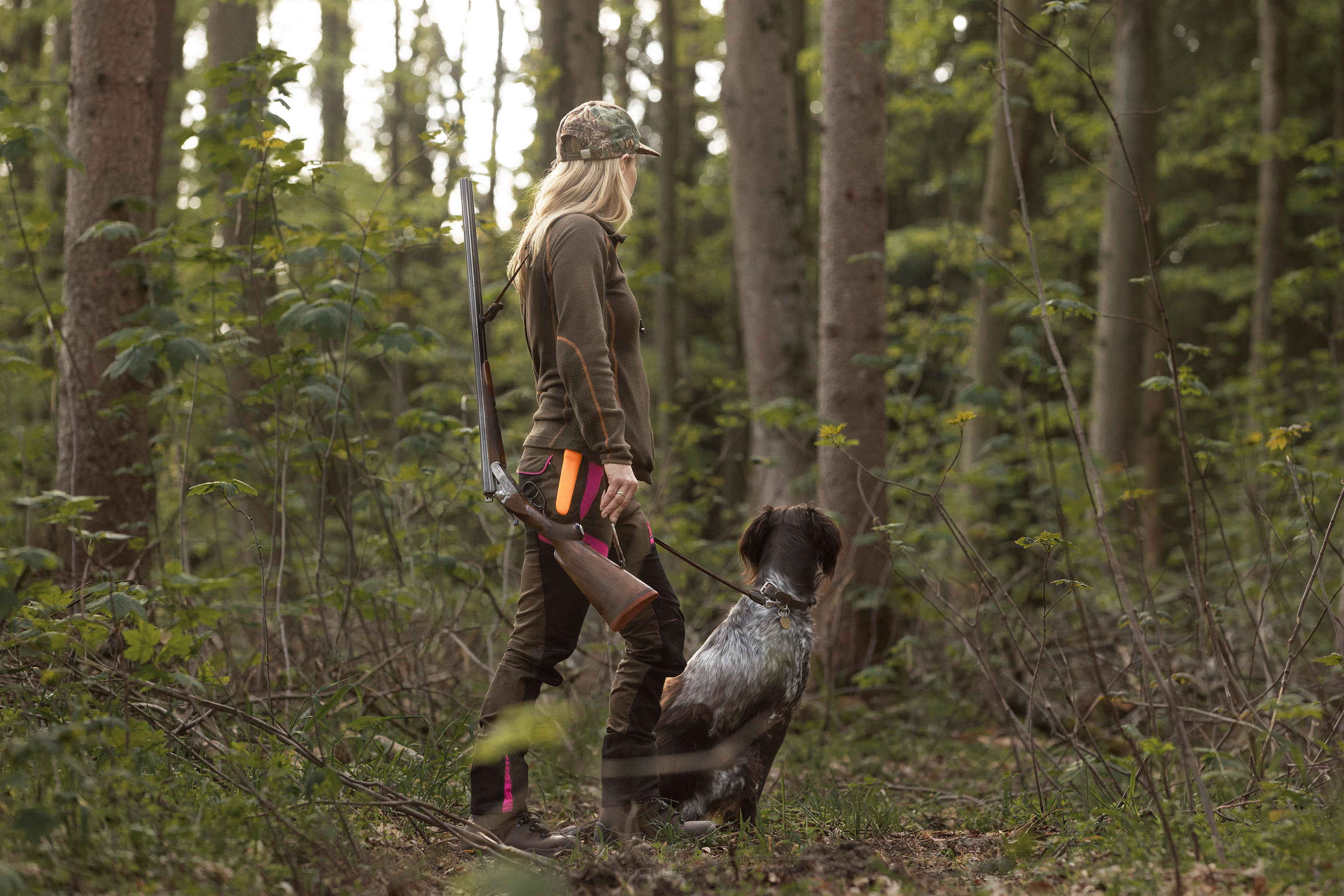 Frau mit gebrochener Flinte über der Schulter und Hund an ihrer Seite blickt in die Ferne