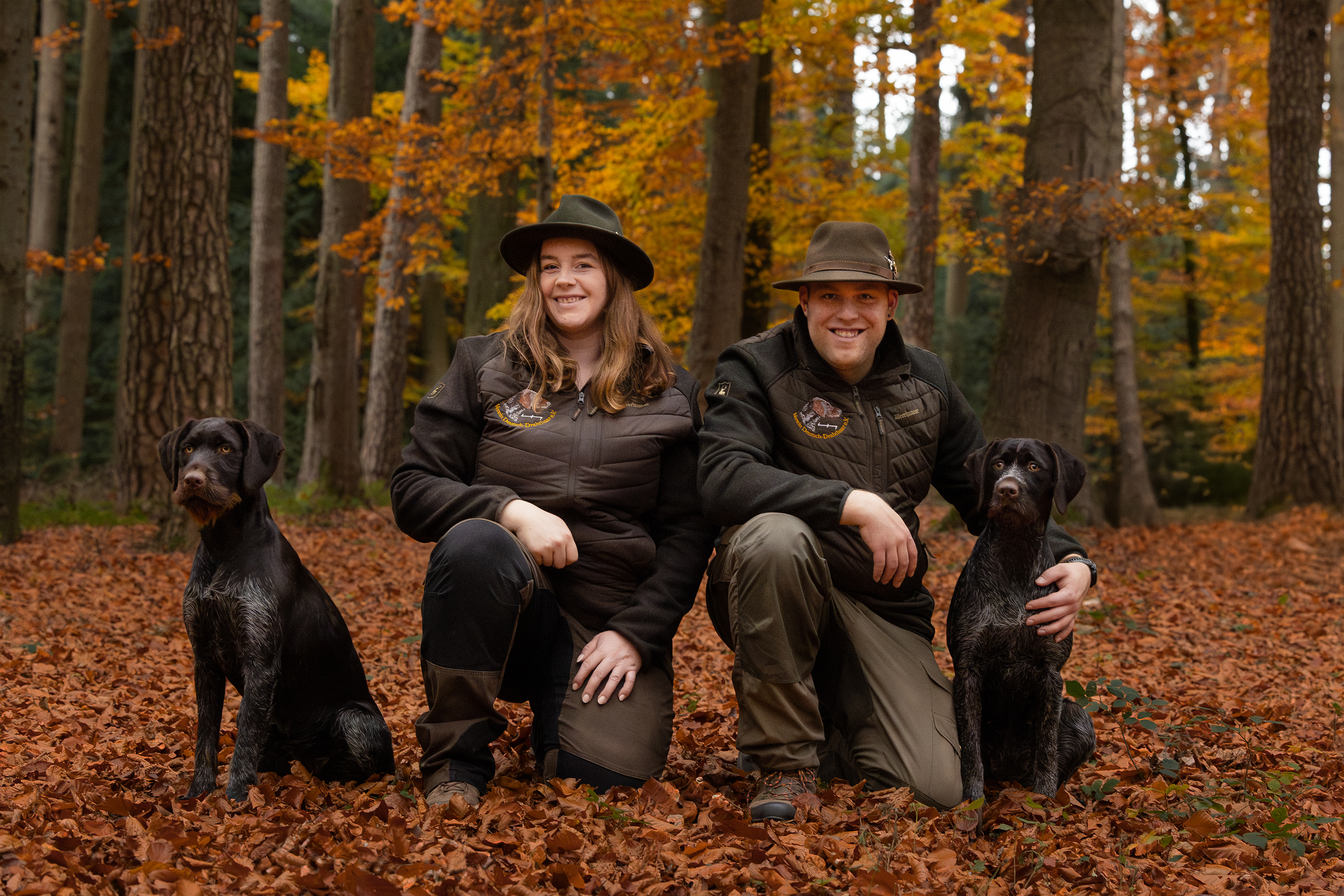 Jägerin und Jäger mit ihren beiden Deutsch Drahthaaren beieinander sitzend